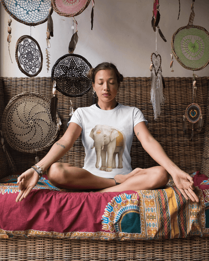 Weißer Elefant Damen T-Shirt auf einer sitzenden Frau in meditativer Pose, umgeben von dekorativen Wandbehängen.