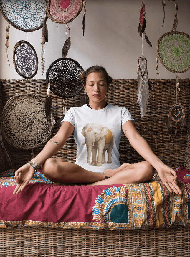 Weißer Elefant Damen T-Shirt auf einer sitzenden Frau in meditativer Pose, umgeben von dekorativen Wandbehängen.